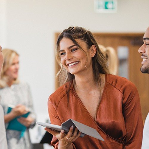 A woman in an orange blouse smiles while holding a tablet, engaging with two people in conversation. A group of individuals stands in the background, indicating a professional setting.