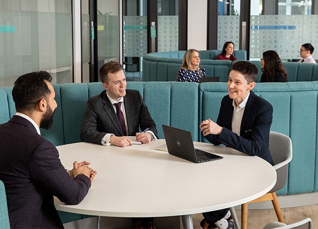 Three professionals are engaged in a discussion around a round table with a laptop. The setting features modern furniture and a relaxed atmosphere, with colleagues visible in the background.
