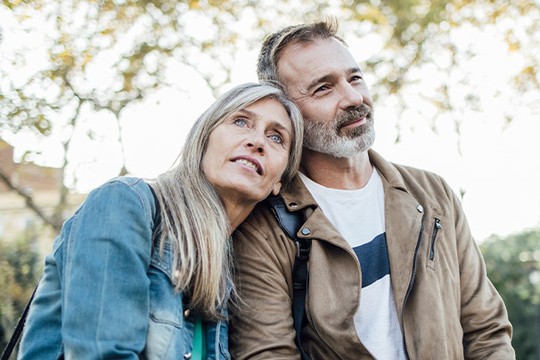 A couple leans close together, with the woman resting her head on the man's shoulder. They appear relaxed and affectionate, against a backdrop of softly blurred trees and greenery.