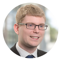 A young individual with short, light hair and glasses smiles while dressed in formal attire. The background features a blurred, professional environment with soft lighting, suggesting a business or corporate setting.
