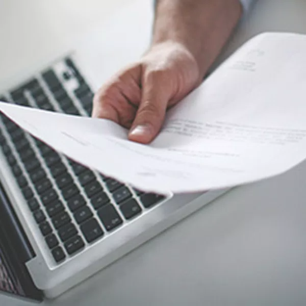 A hand holds a sheet of paper, possibly a document, in front of a laptop keyboard. The setting suggests a workspace or office environment.