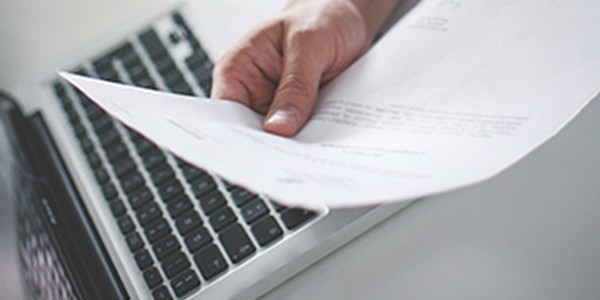 A hand holds a sheet of paper, possibly a document, in front of a laptop keyboard. The setting suggests a workspace or office environment.