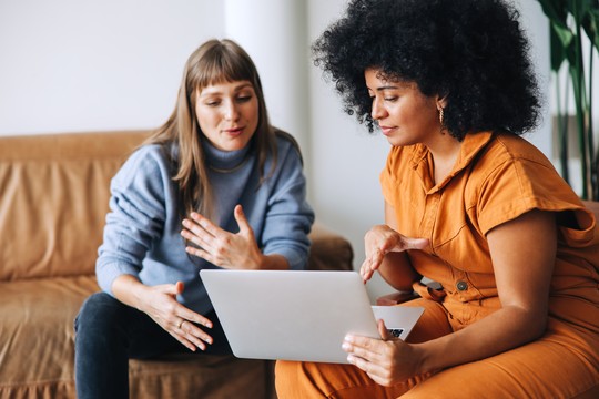 Two women sit on a couch, engaging in conversation. One points at a laptop screen while the other listens attentively, in a bright, modern indoor space with plants in the background.