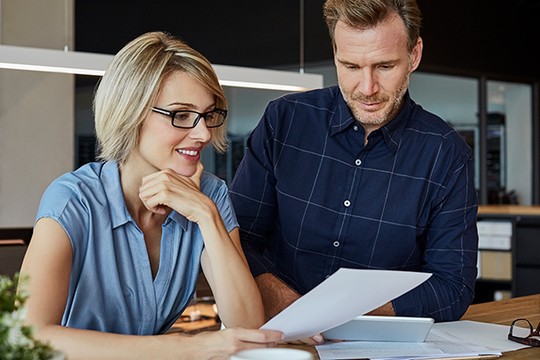 A woman in glasses and a blue shirt reviews a document with a man in a plaid shirt, both engaged in discussion at a modern workspace with natural lighting.