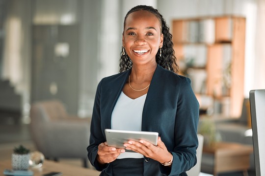 A woman holds a tablet and smiles confidently while standing in a modern office environment with soft lighting, elegant furniture, and greenery in the background, creating a professional atmosphere.