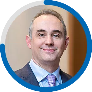 A man with short gray hair smiles confidently. He is wearing a formal suit with a light blue shirt and a pink tie, set against a blurred indoor background.
