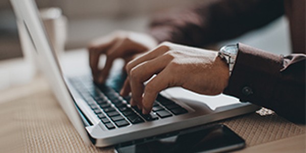 A person is typing on a laptop keyboard, with hands positioned over the keys. The background suggests a workspace, featuring a smartphone and an organized table surface.