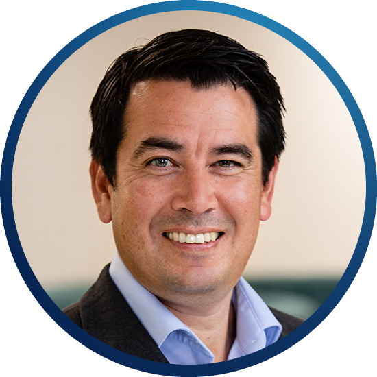 A man with short dark hair smiles warmly, wearing a blue shirt under a dark blazer. He is framed within a circular border against a light background.