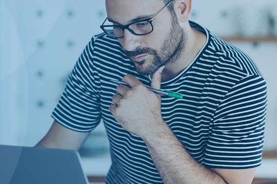 A man with glasses and a striped shirt is focused on a laptop, holding a pen. He is seated in a modern kitchen with light-colored cabinetry and shelves in the background.