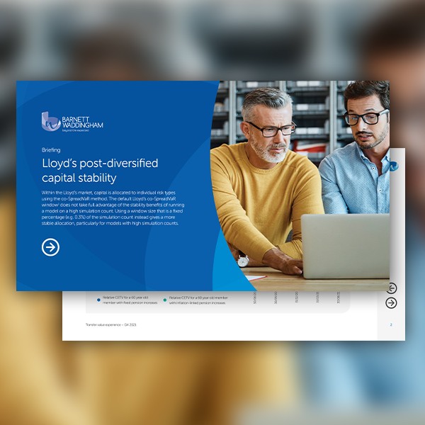 Two men are sitting at a desk, reviewing a document on a laptop. The setting appears to be a modern office, with a blue and white presentation visible on the screen. Text indicates a briefing about "Lloyd's post-diversified capital stability."