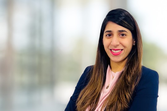 A woman with long, dark hair smiles confidently, wearing a dark blazer over a light blouse. She stands in a softly blurred office environment, conveying professionalism and approachability.