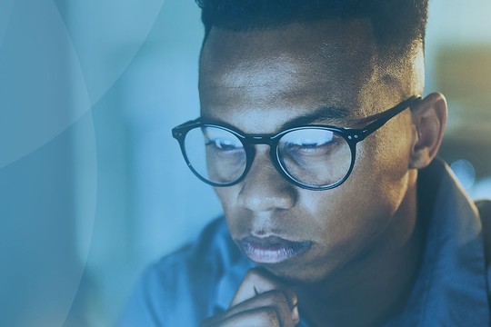 A young man with glasses is focused on a screen, resting his chin on his hand. The background features a warm light, creating a cozy, introspective atmosphere.