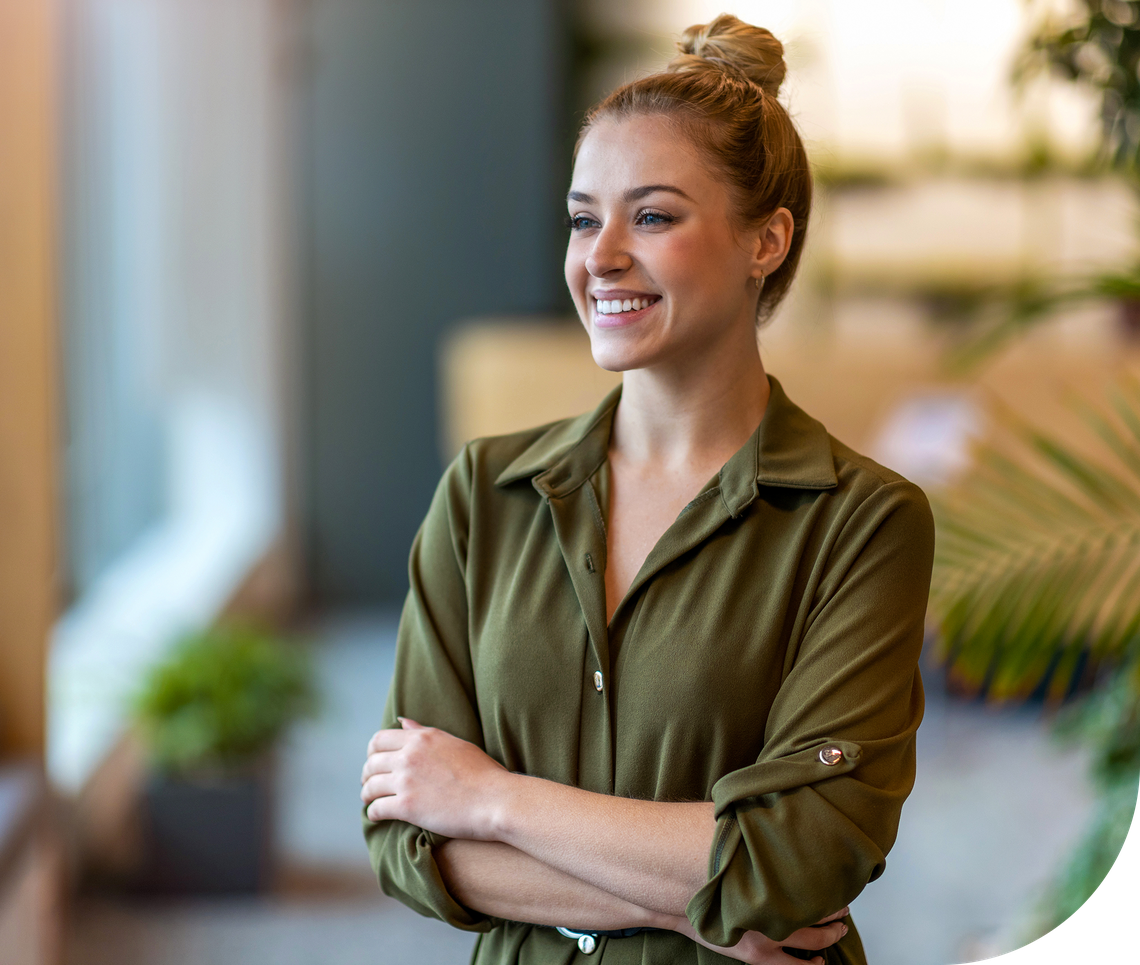 A woman stands with her arms crossed, smiling warmly. She wears an olive green shirt and is in a bright, modern indoor space with plants and soft furniture in the background.