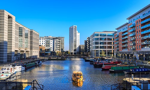 A yellow boat navigates through a calm canal surrounded by modern buildings. The clear blue sky reflects on the water, enhancing the urban scenery. Several moored boats line the edges.
