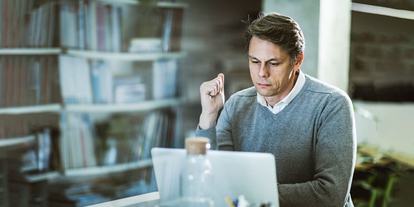 A man sits at a desk, focusing on his laptop. He raises his hand thoughtfully, surrounded by bookshelves, with green plants visible in the bright, modern workspace.