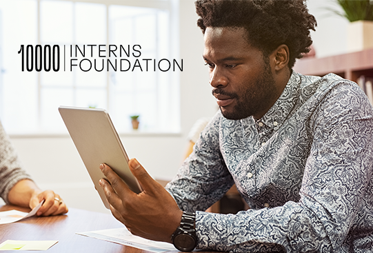 A young man sits at a wooden table, intently holding a tablet while engaged in work. Papers are scattered around him in a bright, modern office space. Text above reads: "10000 INTERNS FOUNDATION."