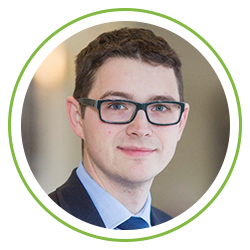 A man with short dark hair and glasses smiles confidently, dressed in a suit and blue shirt. He is framed within a circular green border, suggesting a professional or formal context.