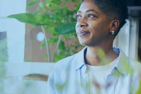 A young person with short hair gazes thoughtfully to the side, surrounded by greenery, in a softly lit indoor space with light blue tones in the background.