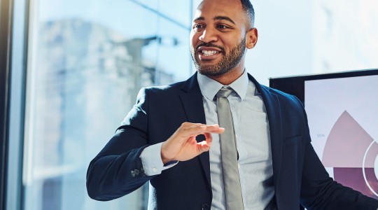 A man in a suit presents information using a screen displaying graphs and text, engaging with an audience in a bright office setting with large windows.