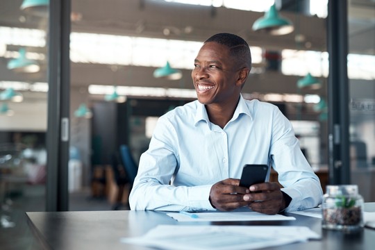 A man in a light blue shirt smiles while holding a smartphone. He is seated at a table with documents, in a modern workspace filled with natural light and green pendant lights.