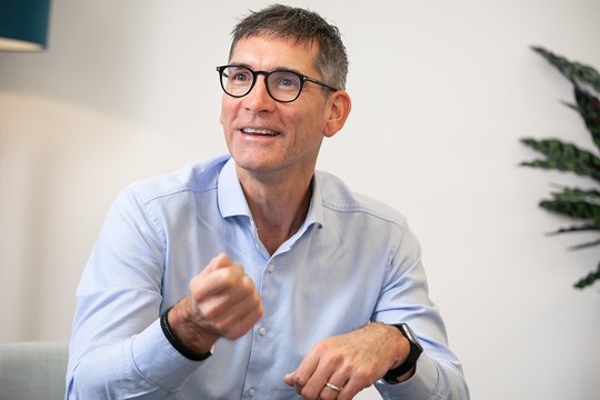 A man with glasses is smiling and gesturing with one hand while sitting in a light, modern room with a plant in the background, indicating a friendly discussion.