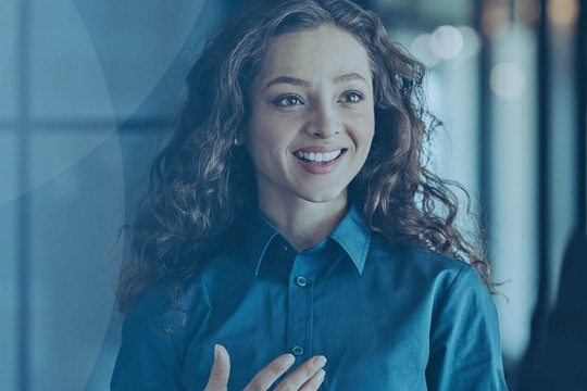 A woman with curly hair wears a blue shirt and smiles while gesturing with her hand, interacting with a person dressed in a suit in a modern, softly-lit environment.