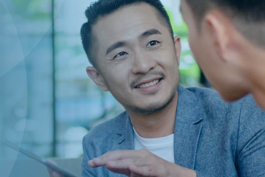 Two men are engaged in a conversation, with one gesturing toward a tablet. They are in a bright, modern space with greenery visible in the background.