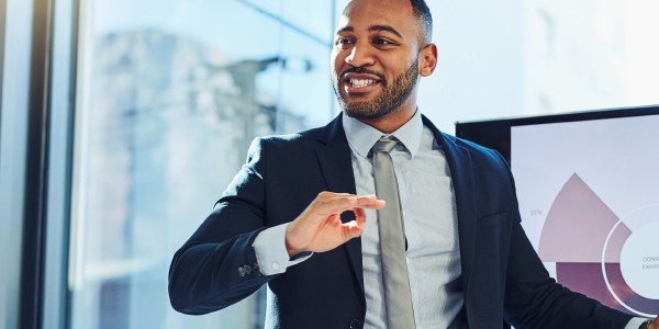 A man in a suit presents information using a digital display while engaging with an audience in a well-lit office environment, with large windows and modern decor.