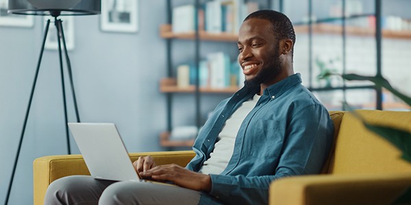 A man sits comfortably on a yellow couch, smiling while using a laptop. The bright, modern living space features bookshelves and plants in the background.