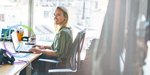 A woman sits at a desk, typing on a laptop, smiling. The workspace features office supplies and a cityscape visible through large windows, creating a bright, modern environment.