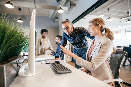 A woman seated at a desk interacts with a man standing beside her, pointing towards a computer screen. Two other individuals are engaged in conversation in the background of a modern office space.