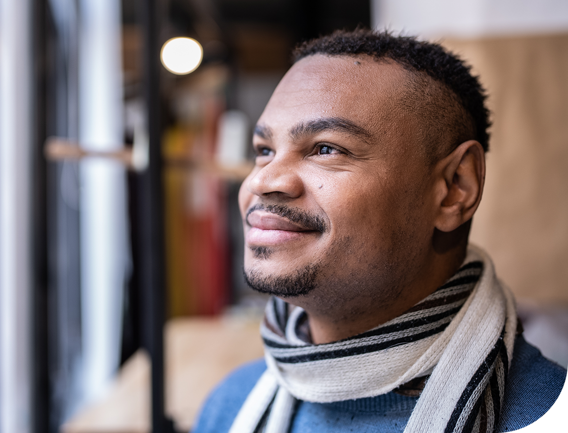A man, wearing a striped scarf, smiles thoughtfully while looking out of a window. The background features warm, blurred colors hinting at a cozy indoor environment.