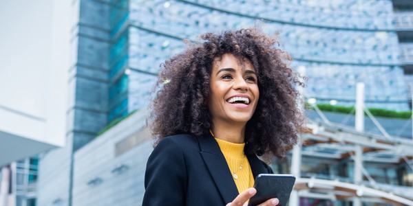 A woman with curly hair smiles while holding a smartphone. She appears joyful in a modern urban setting with glass buildings in the background.
