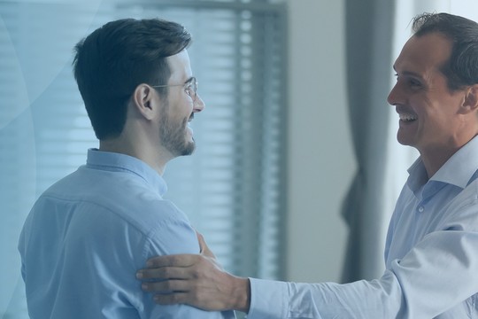 Two men, one with glasses and a light blue shirt, engage in a friendly embrace, smiling. They are in a well-lit, professional interior with window blinds in the background.