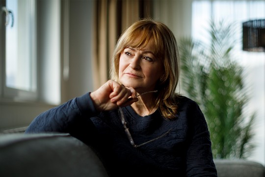 A woman sits pensively on a couch, holding glasses in her hand. Soft natural light floods the room, highlighting a plant and nearby window. She appears contemplative in a cozy setting.