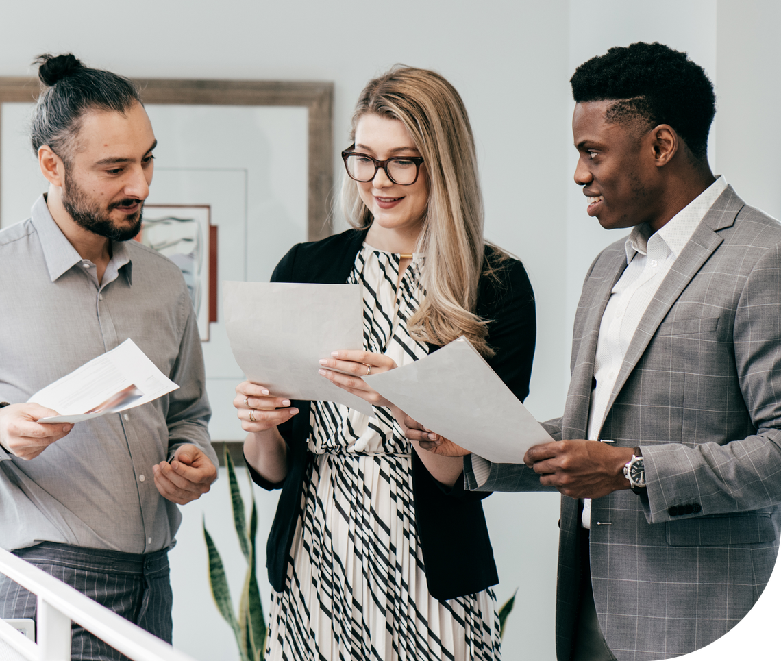 Three colleagues are engaged in discussion while reviewing documents in a modern office setting. They appear focused and collaborative. The environment features light colors and greenery.