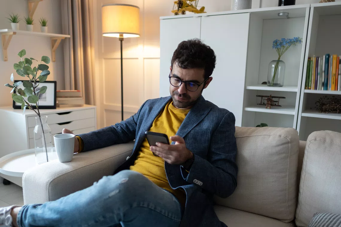 A man sits on a sofa, holding a phone in one hand and a white mug in the other. He wears a blazer over a yellow sweater in a well-lit, cozy living room with decorative plants and books.