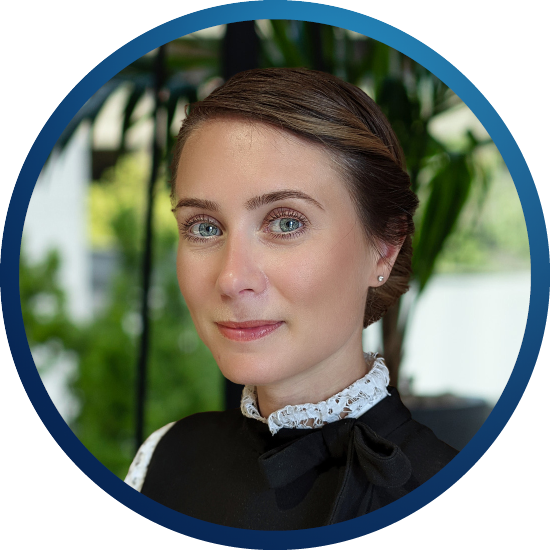 A woman with light brown hair, wearing a black outfit with a white collar and bow, is smiling softly. Green foliage is softly blurred in the background.