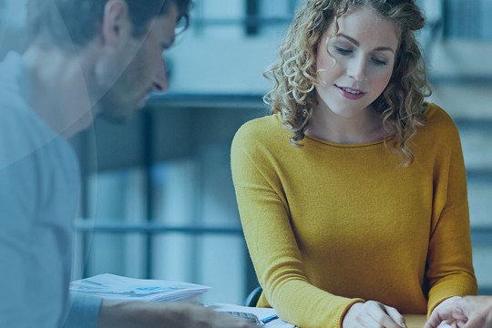 A woman with curly hair in a yellow sweater discusses documents on a table with two men, one in a blue shirt. They appear engaged in conversation in a modern indoor setting with stairs.