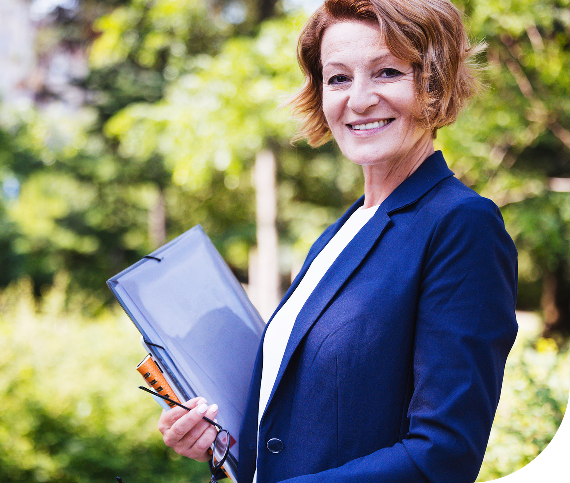 A woman in a blue blazer stands outside, smiling while holding a clipboard and eyeglasses. The background features greenery, indicating a natural, sunny environment.