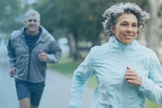 A woman in a light blue jacket jogs along a park path, smiling and engaged in exercise. A man in a gray jacket runs behind her, surrounded by trees and benches.