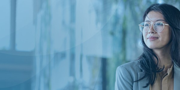 A woman wearing glasses stands confidently, looking to the side. She is in a modern, bright indoor setting with greenery and glass elements in the background.