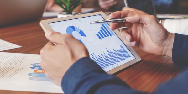 A person holds a tablet displaying graphs and charts, analyzing data during a meeting. The setting includes laptops and paperwork on a wooden table, with a small plant nearby.
