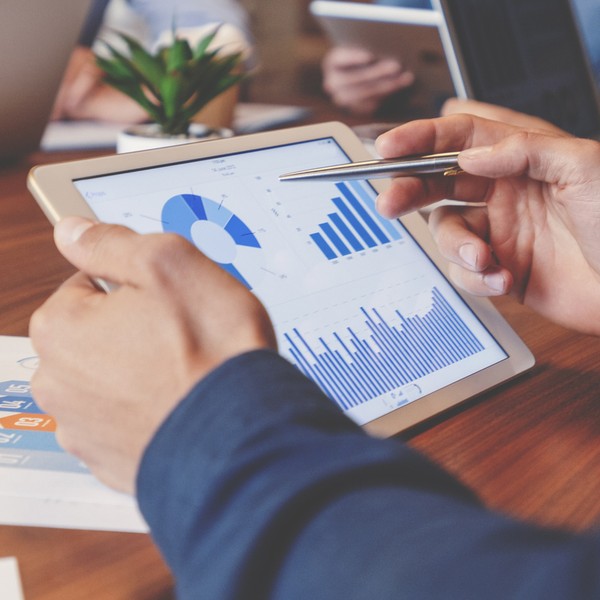 A person holds a tablet displaying graphs and charts, analyzing data during a meeting. The setting includes laptops and paperwork on a wooden table, with a small plant nearby.