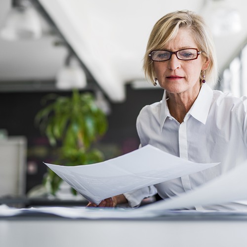 A middle-aged woman, wearing glasses and a white shirt, meticulously reviews documents at a desk in a modern office with natural light and green plants in the background.