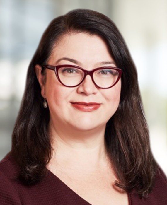 A woman with long dark hair is smiling, wearing glasses and a burgundy top. The blurred background suggests a professional indoor setting, possibly an office or conference area.
