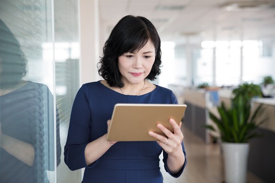 A woman in a blue dress is holding a tablet, focused on its screen. She stands near a glass wall in a modern office filled with plants and workstations.
