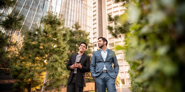 Two men, one in a black suit and the other in a blue suit, walk and smile along a pathway surrounded by greenery and modern buildings, enjoying a conversation.