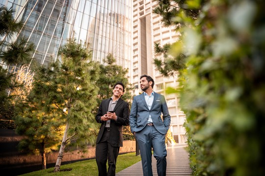Two men, one in a black suit and the other in a blue suit, walk and smile along a pathway surrounded by greenery and modern buildings, enjoying a conversation.