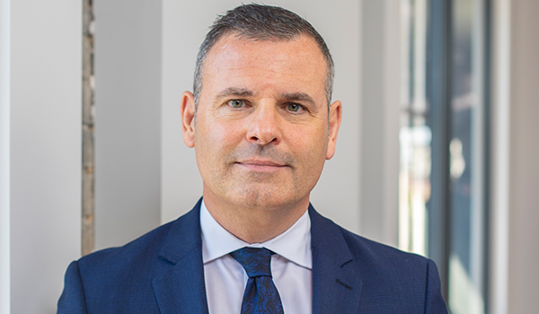 A man in a navy suit stands confidently, facing the camera. He has short, neatly styled hair and a slight smile, positioned against a bright, modern indoor setting with large windows.